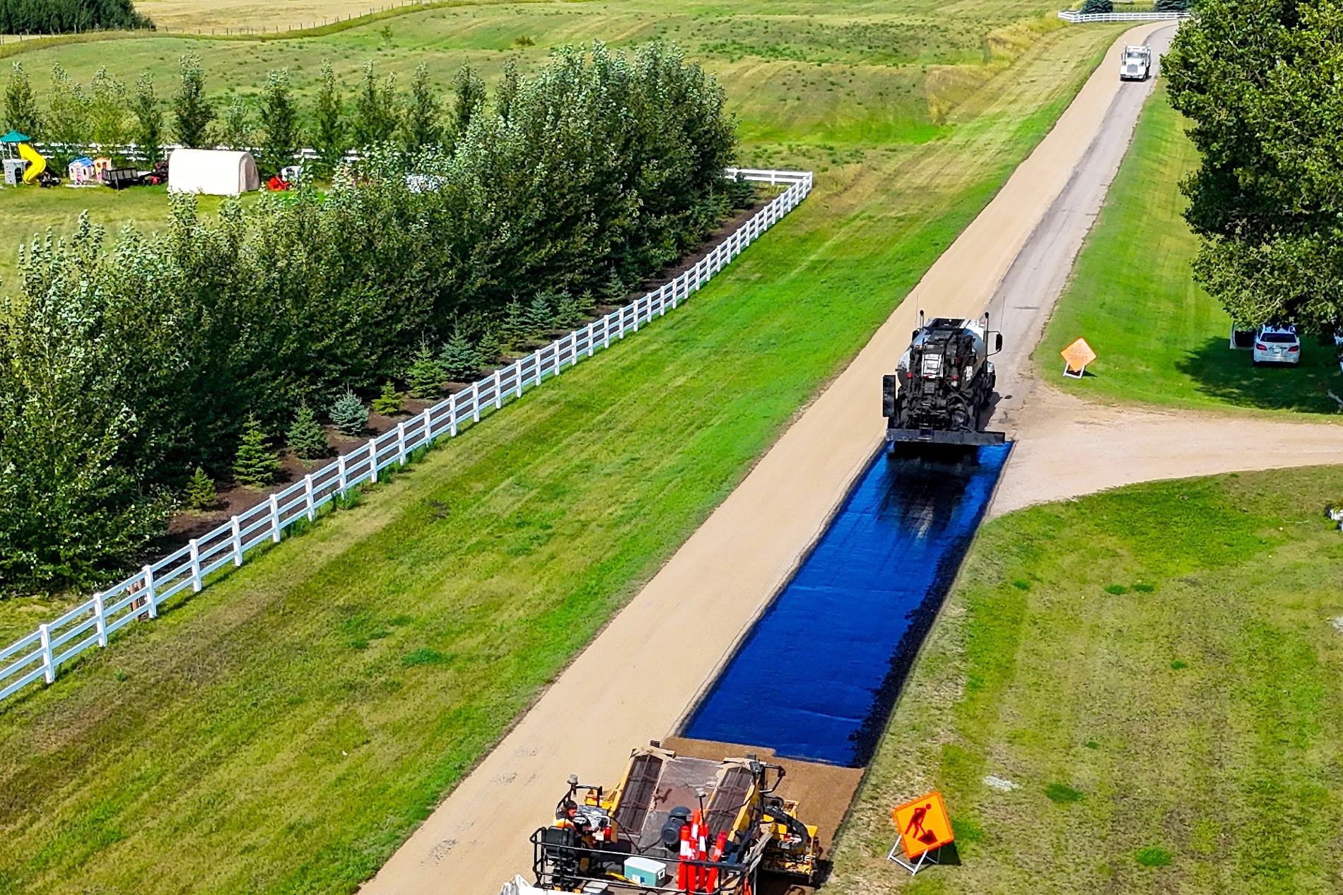 The same driveway a few metres on: fresh black hot mix asphalt laid between the white fence and the field